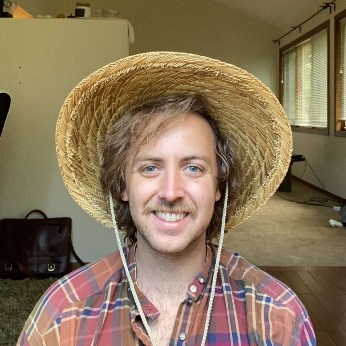 Sam Holtzen wearing a straw sunhat and a plaid shirt, smiling.